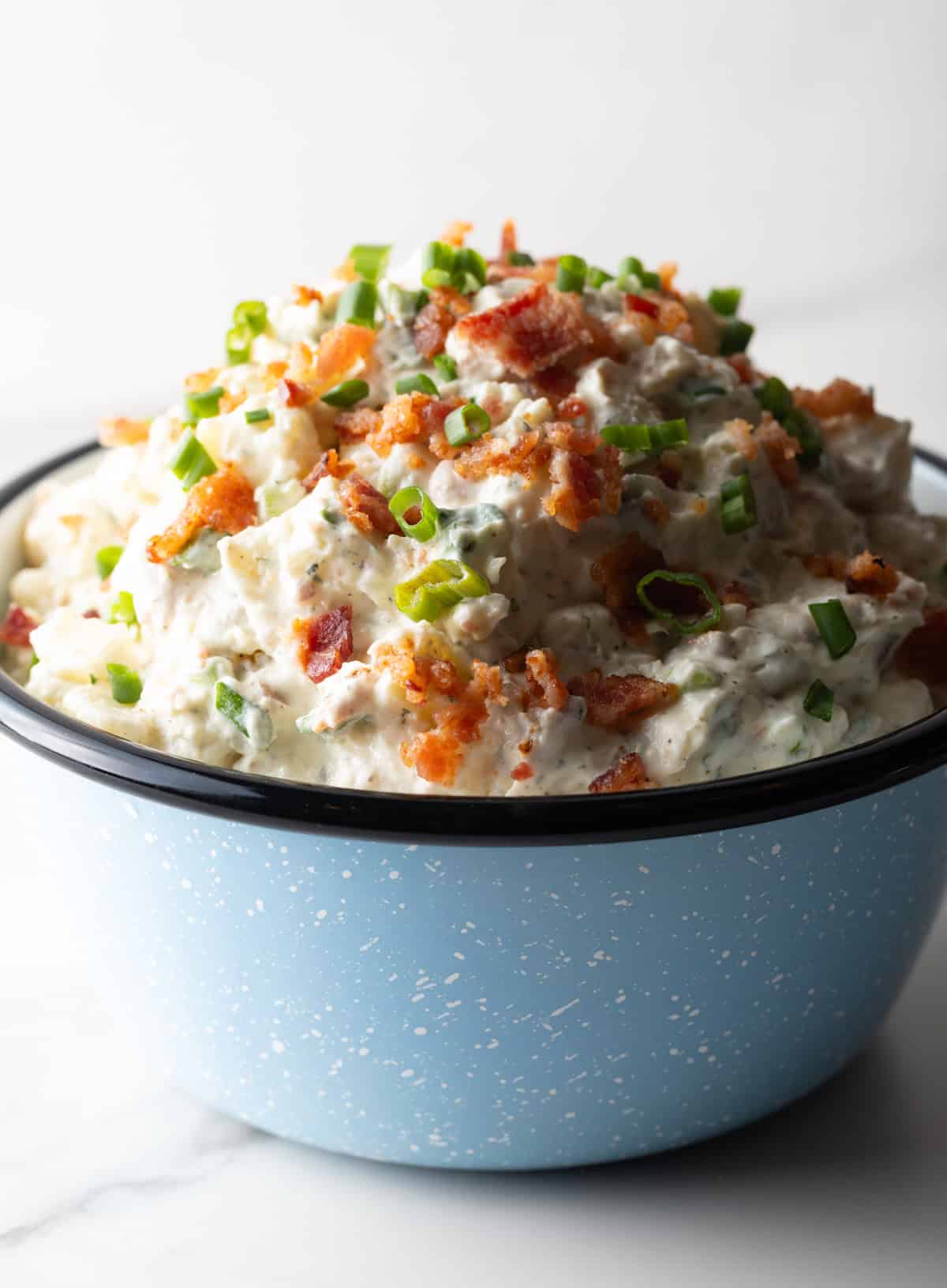A blue bowl filled with a bacon ranch potato salad recipe topped with green onion and chopped bacon.