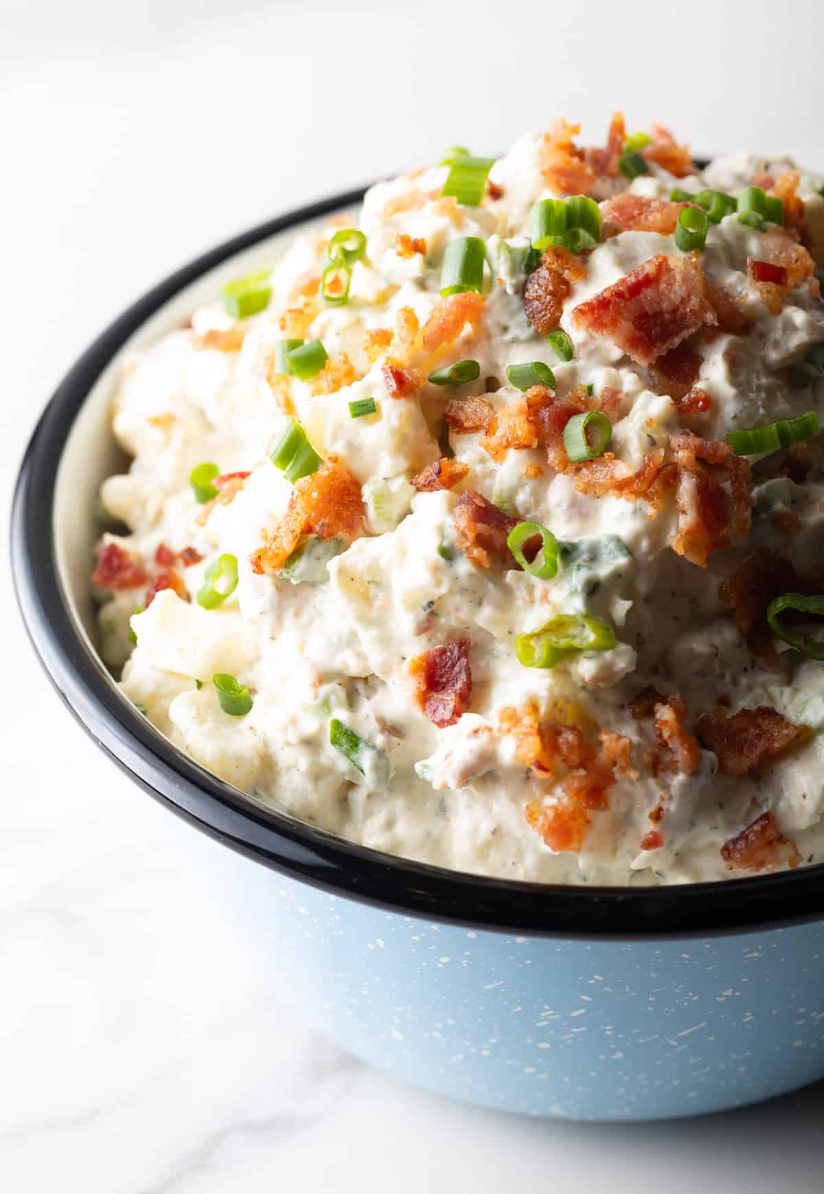 A blue bowl filled with bacon ranch potato salad topped with green onion and chopped bacon.