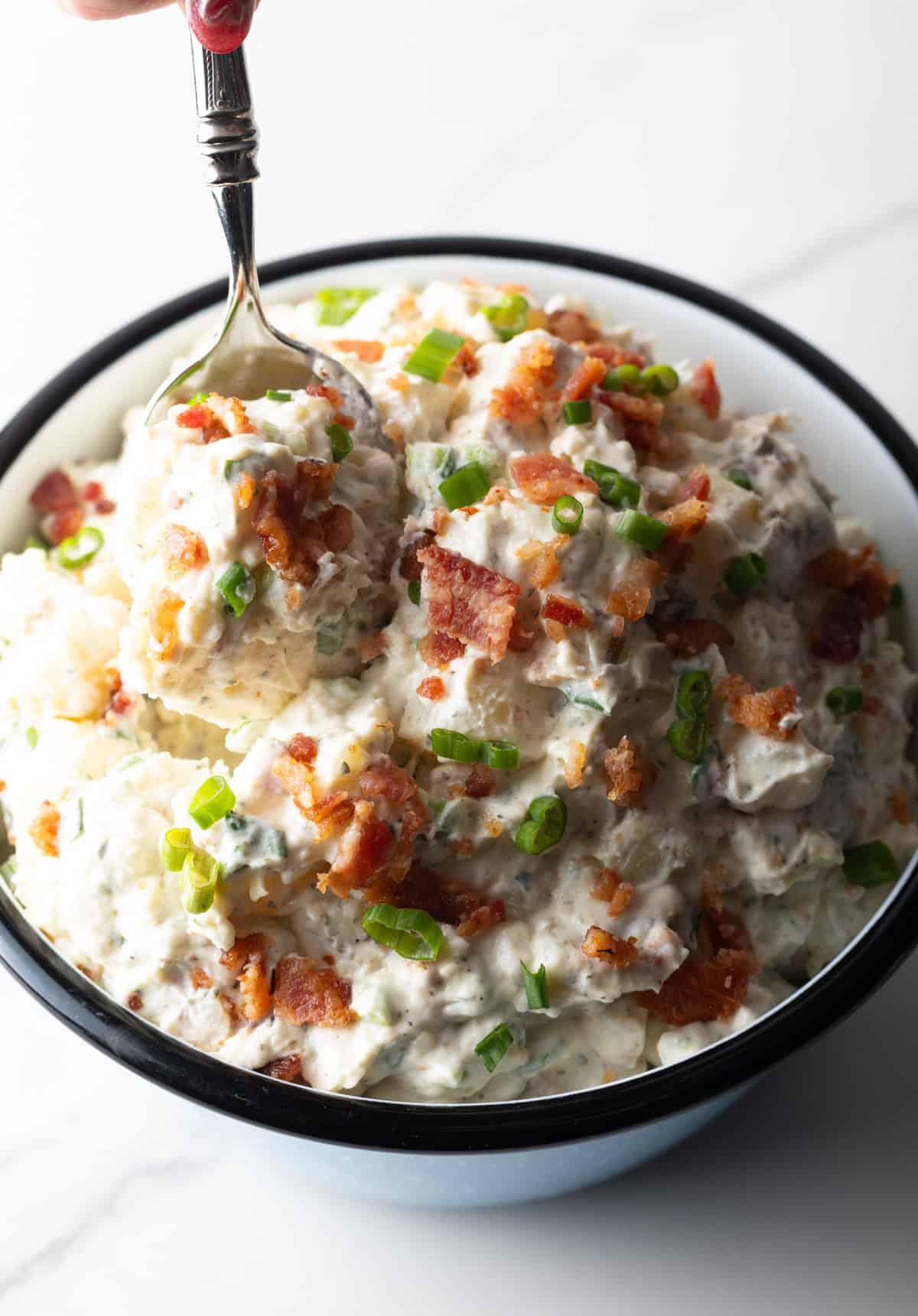 Bacon ranch potato salad is being mixed with a metal spoon in a bowl.
