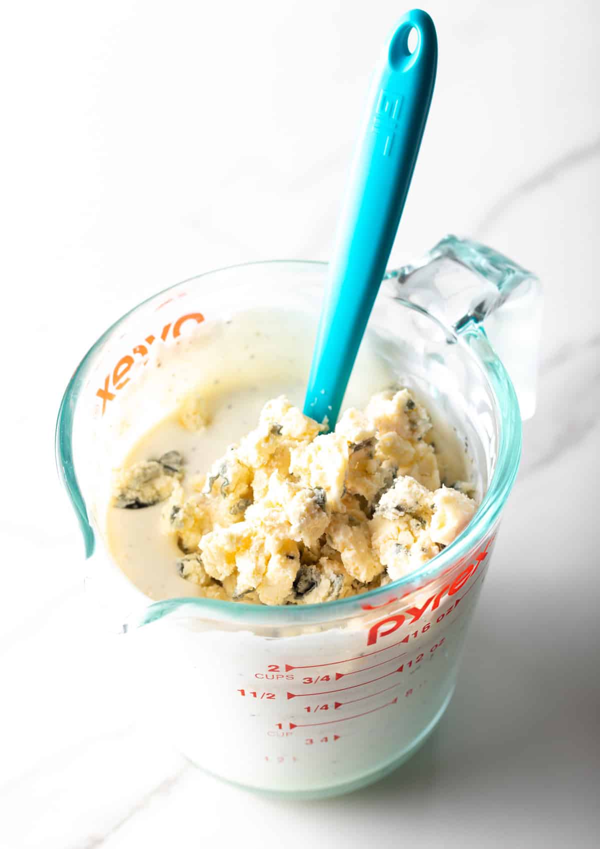 Blue cheese crumbles are being added to a glass measuring bowl for a blue cheese dressing recipe.