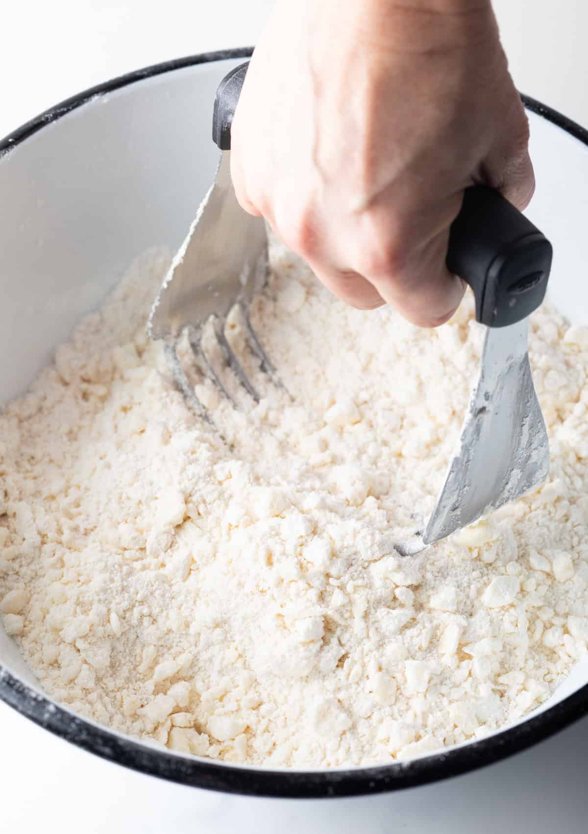 A hand holding a pastry cutter is mixing butter and flour together in a bowl.