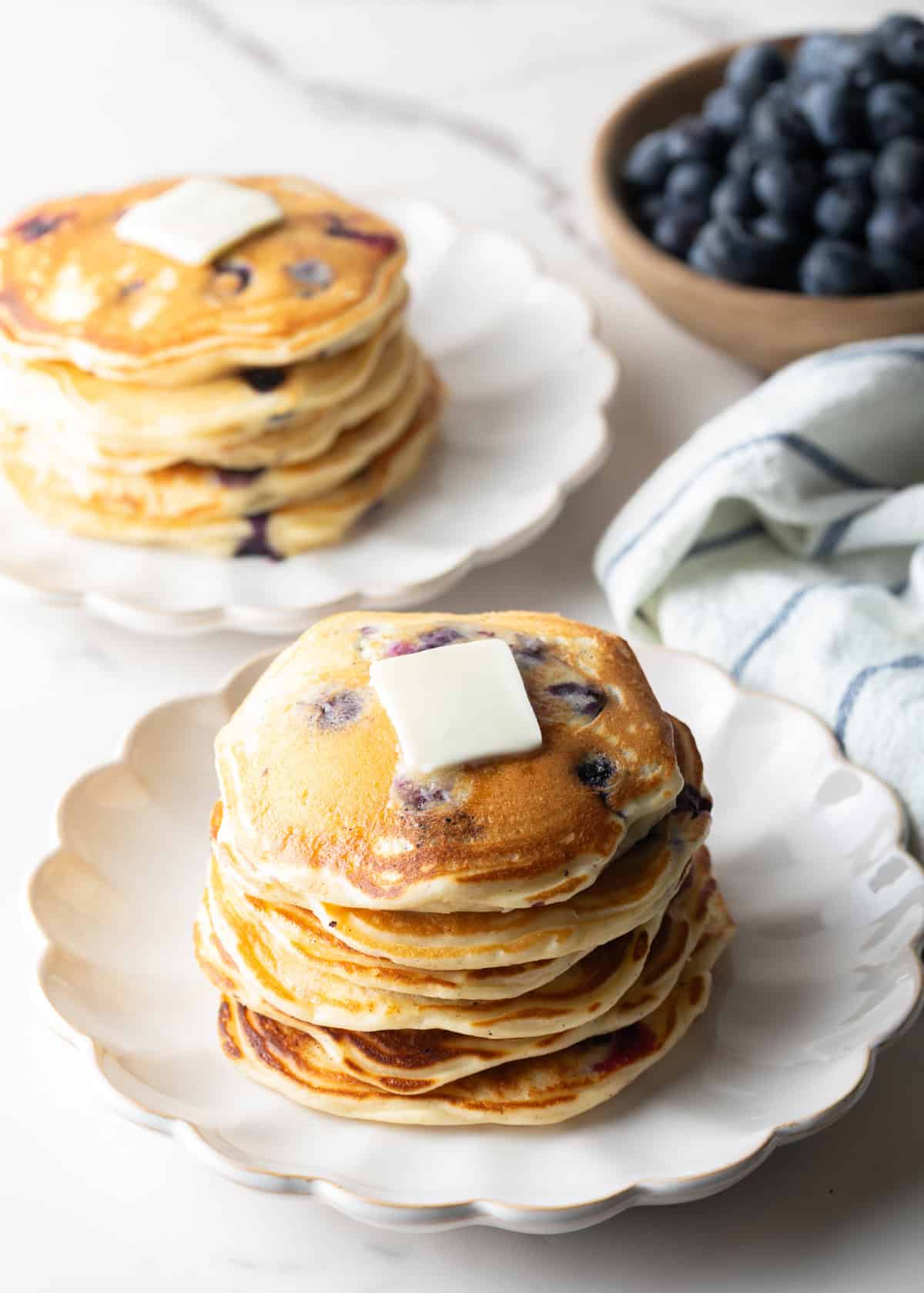 A large stack of blueberry pancakes in the foreground, another stack of pancakes in the background with a bowl of fresh berries. The stack closest to camera has a pat of butter on top.