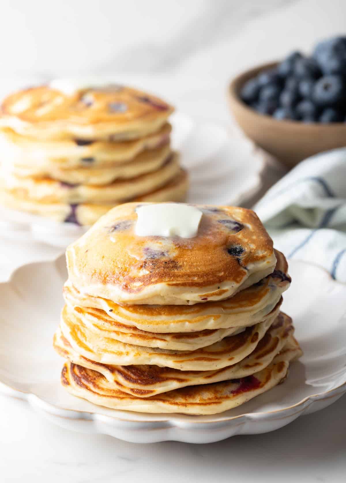 A large stack of blueberry pancakes in the foreground, another stack of pancakes in the background with a bowl of fresh berries. The stack closest to camera has a pat of butter on top.