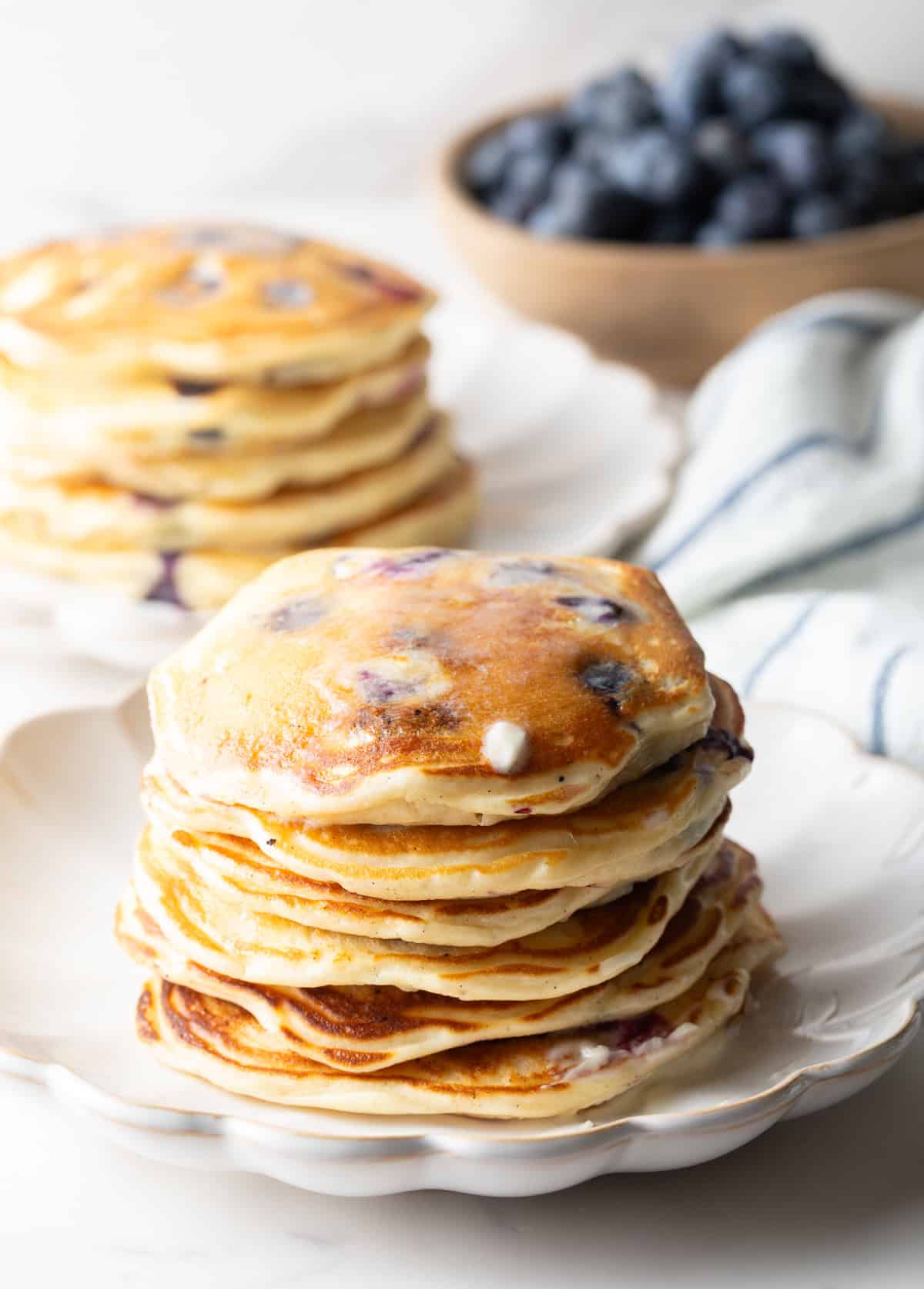 A large stack of blueberry pancakes in the foreground, another stack of pancakes in the background with a bowl of fresh berries.
