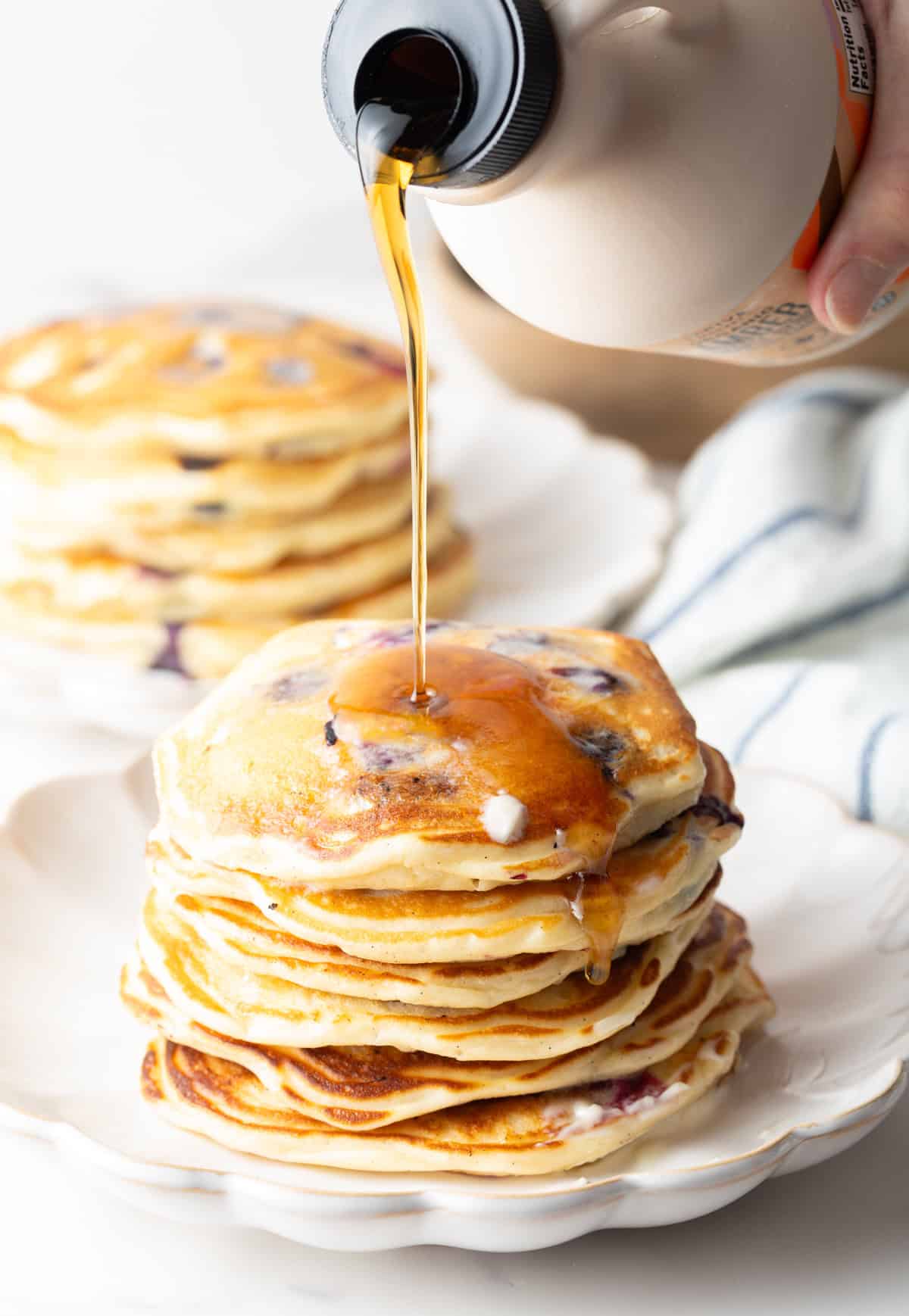 A large stack of pancakes on a white plate, being coated in a drizzle of syrup.