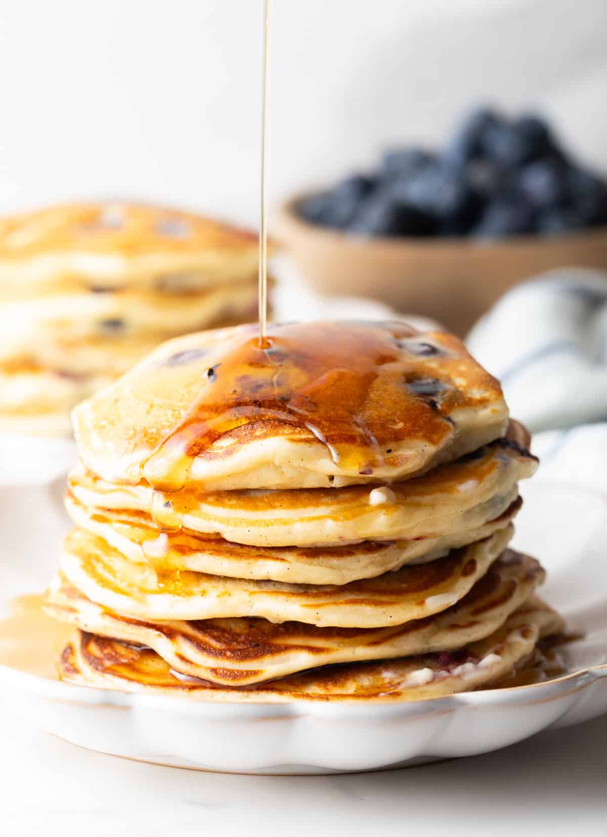 A large stack of pancakes on a white plate, being coated in a drizzle of syrup.