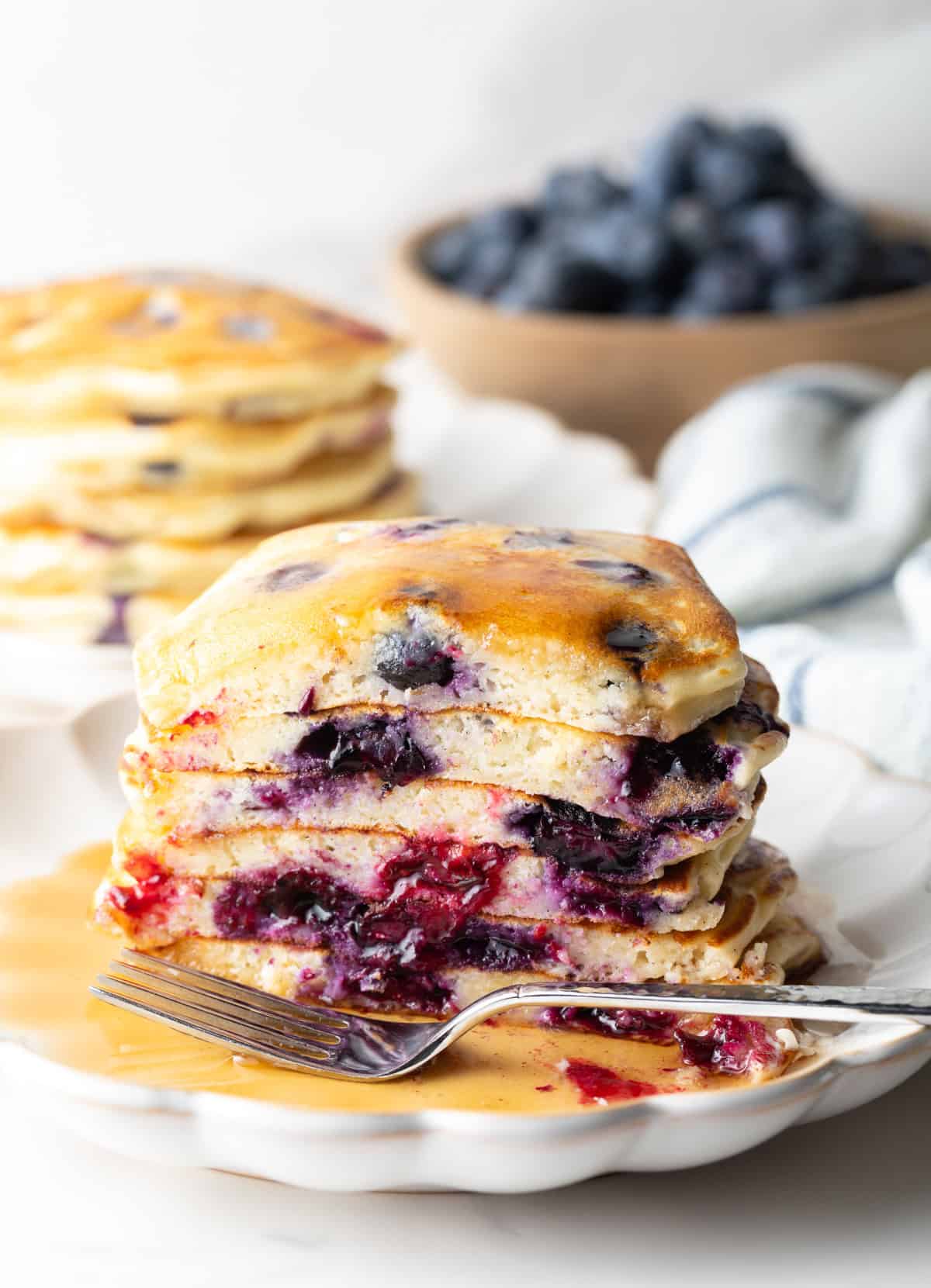 Stack of several blueberry pancakes on a plate, close up view of the pancakes cut in half to show the berries inside. There is a light puddle of syrup and fork on the plate.