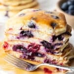 Stack of several blueberry pancakes on a plate, close up view of the pancakes cut in half to show the berries inside. There is a light puddle of syrup and fork on the plate.