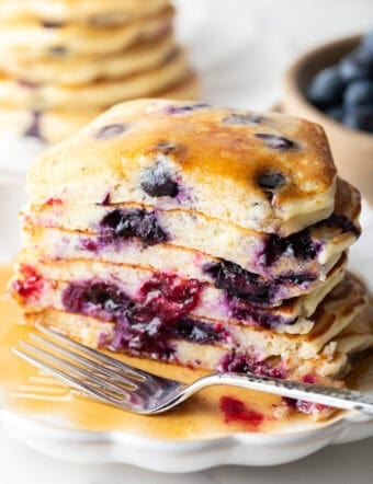 Stack of several blueberry pancakes on a plate, close up view of the pancakes cut in half to show the berries inside. There is a light puddle of syrup and fork on the plate.