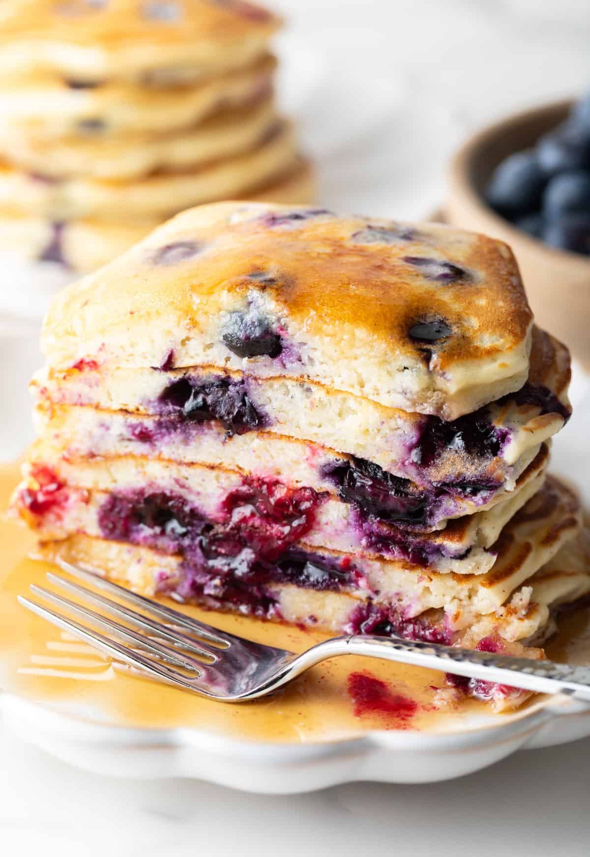 Stack of several blueberry pancakes on a plate, close up view of the pancakes cut in half to show the berries inside. There is a light puddle of syrup and fork on the plate.