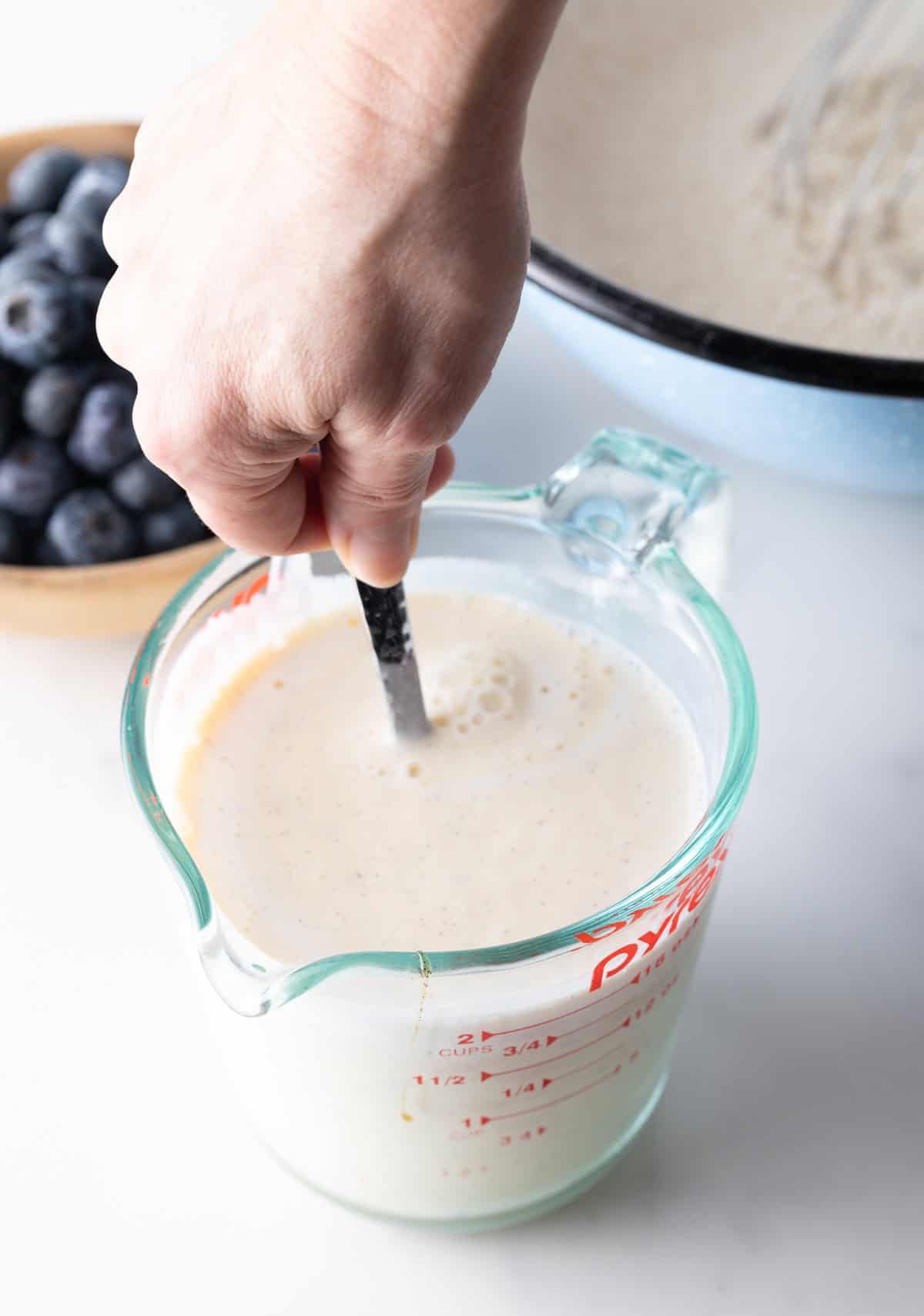 Hand with a metal spoon stirring wet batter ingredients together in a large glass measuring bowl.