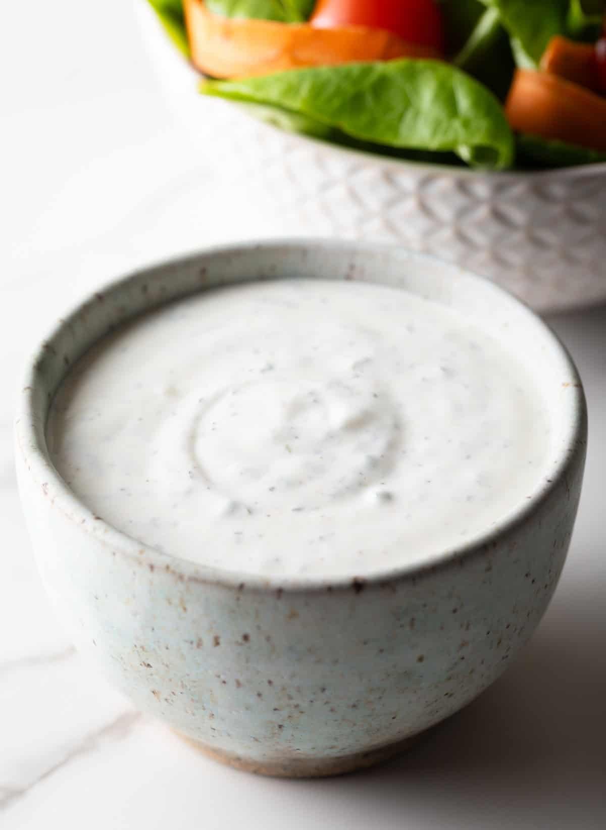A homemade buttermilk ranch dressing recipe in a white bowl, sitting next to a fresh greens salad.