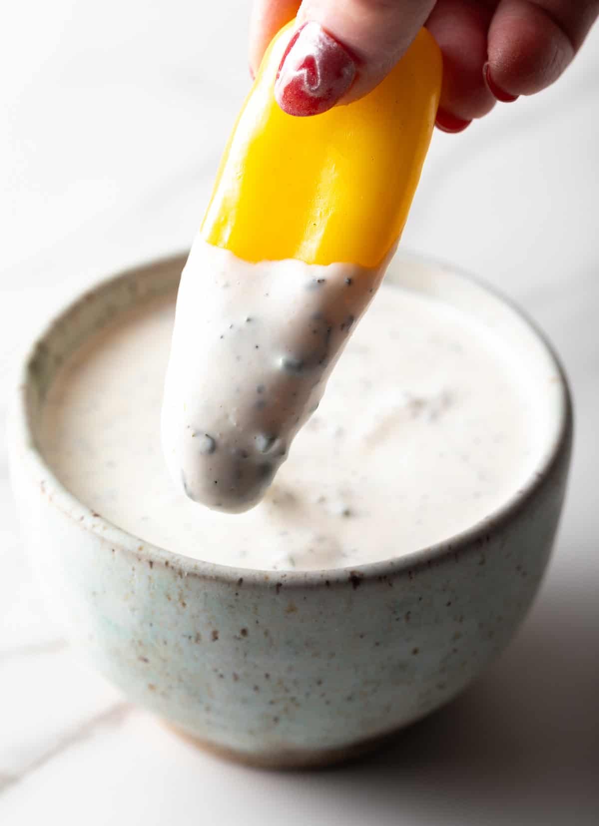 A yellow pepper is being dipped into a bowl filled with homemade ranch dressing made with buttermilk.