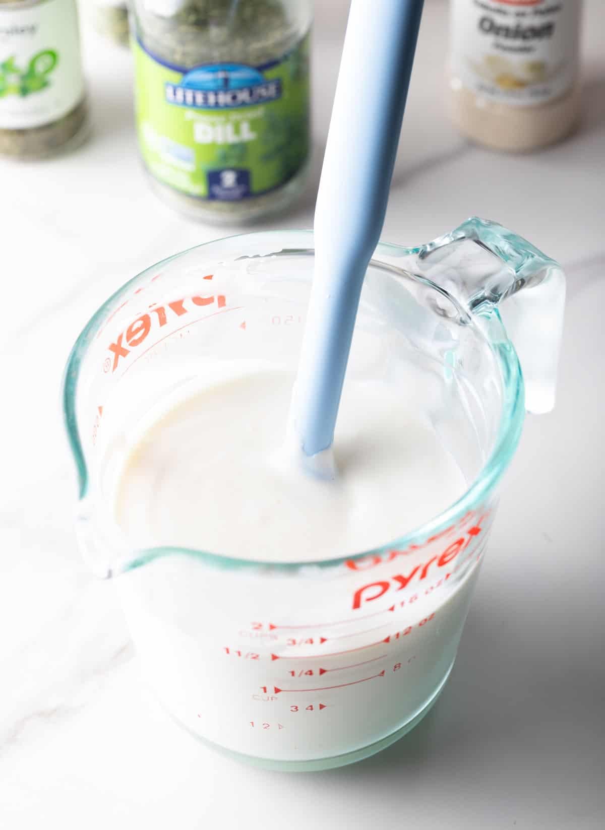 A glass measuring pitcher filled with the creamy base of a buttermilk ranch recipe, being stirred by a spatula.