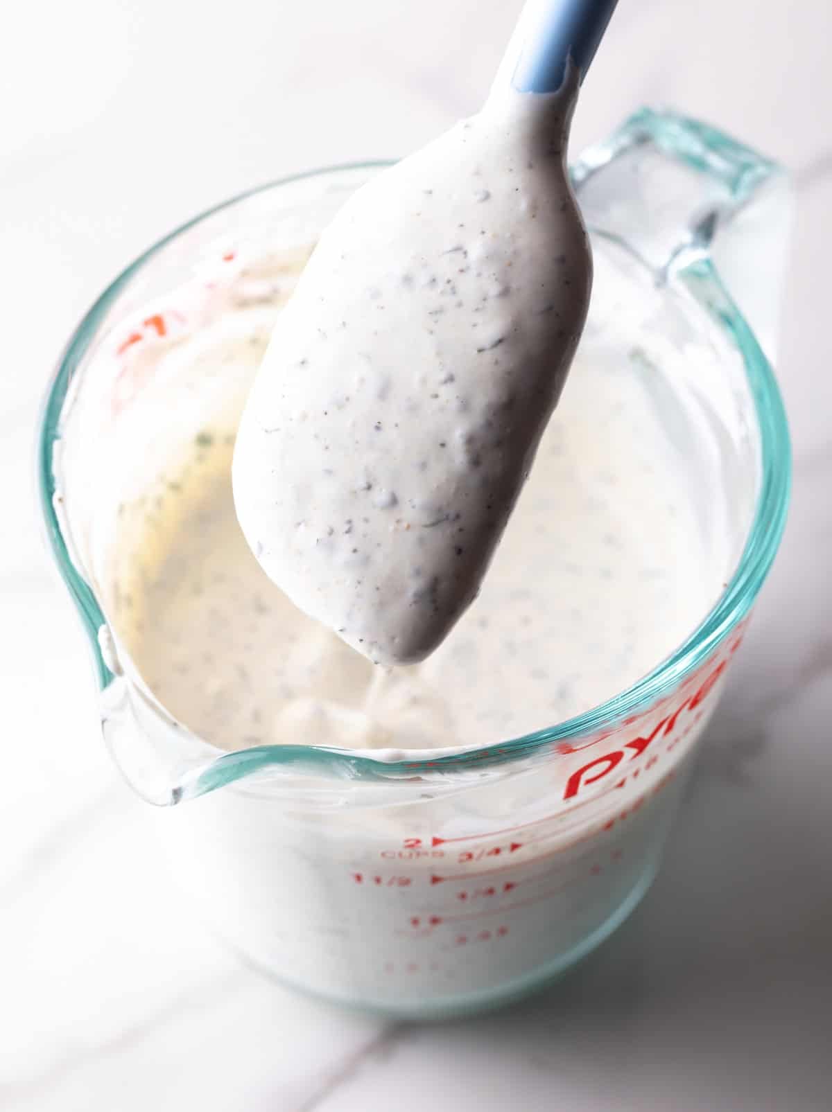 A spatula holding freshly made homemade buttermilk ranch dressing above a glass measuring pitcher filled with the dressing.