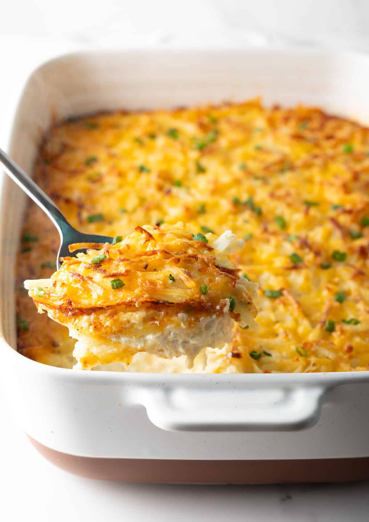 A spatula is taking a scoop out of a baking dish filled with a cheesy hash brown casserole.