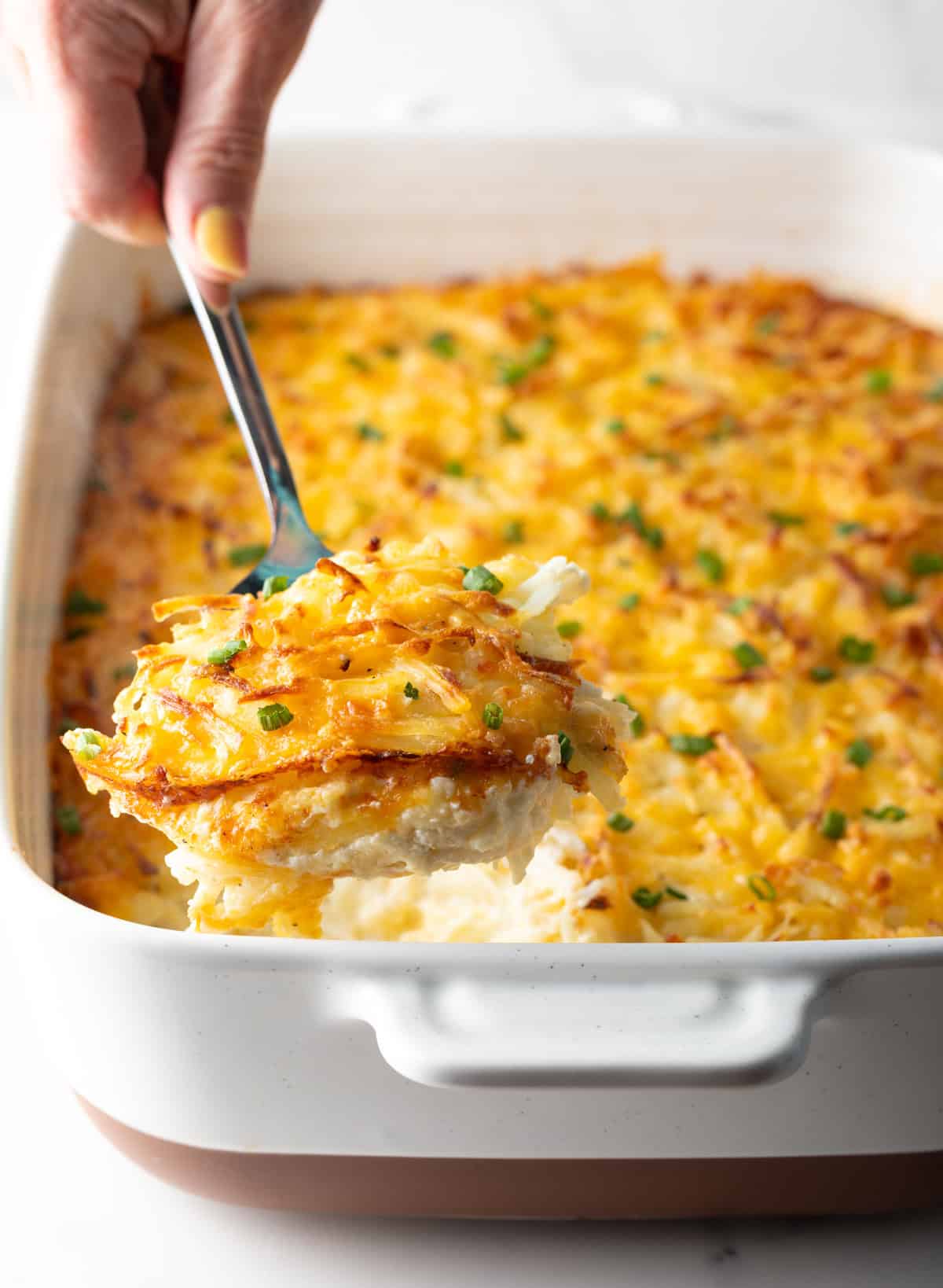 A spatula is taking a scoop out of a baking dish filled with a cheesy hash brown casserole.