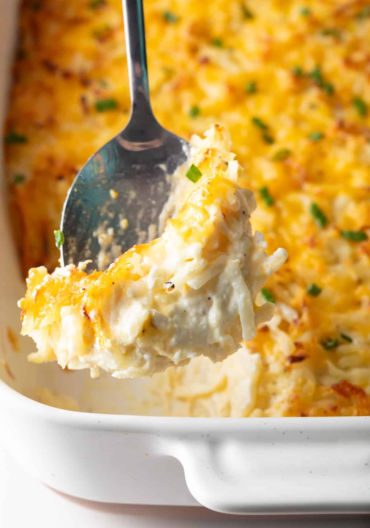 A scoop of cheesy hash brown potato casserole is being held above the casserole dish.