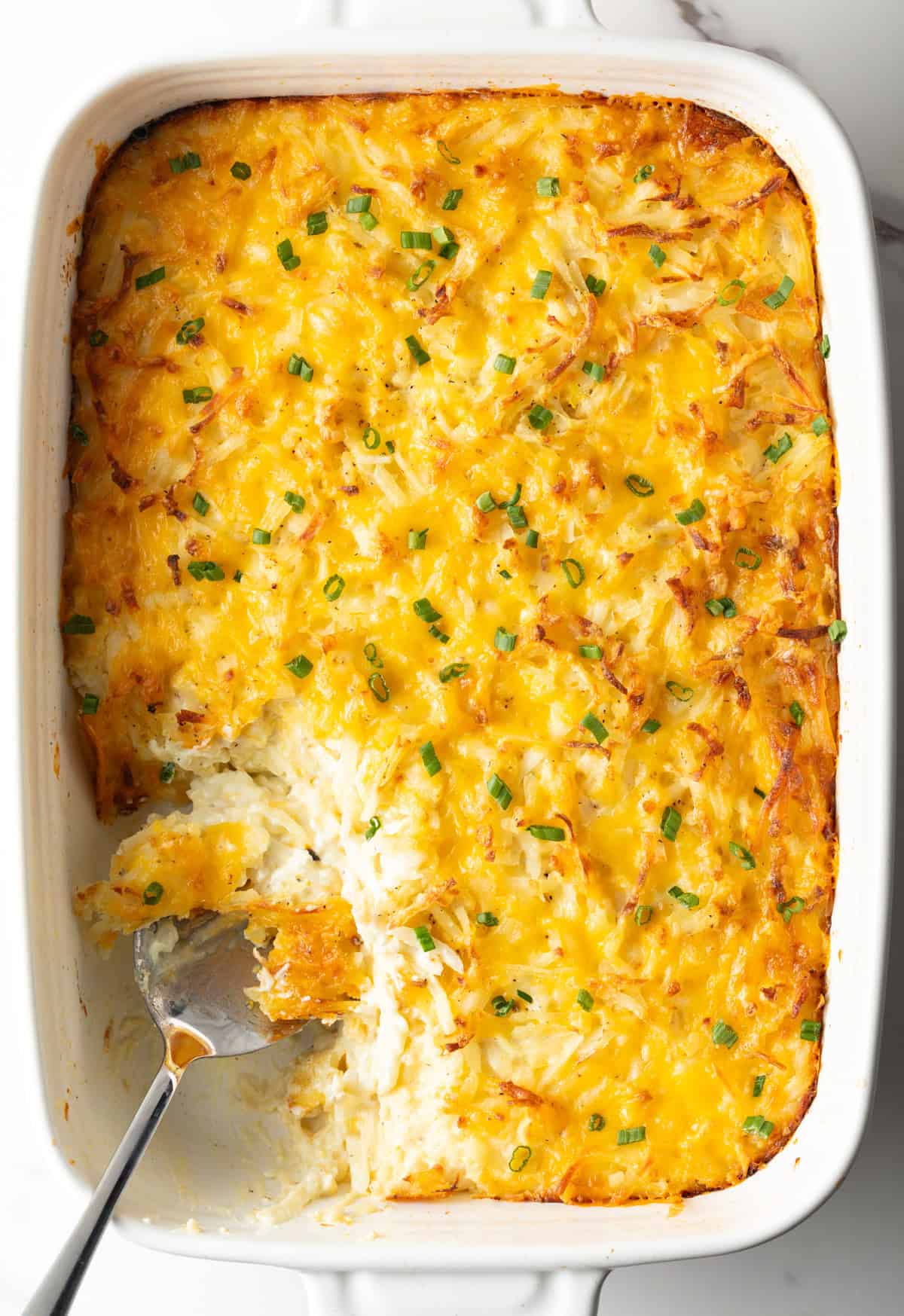 A baking tray filled with cheesy hash brown casserole with a scoop taken out.