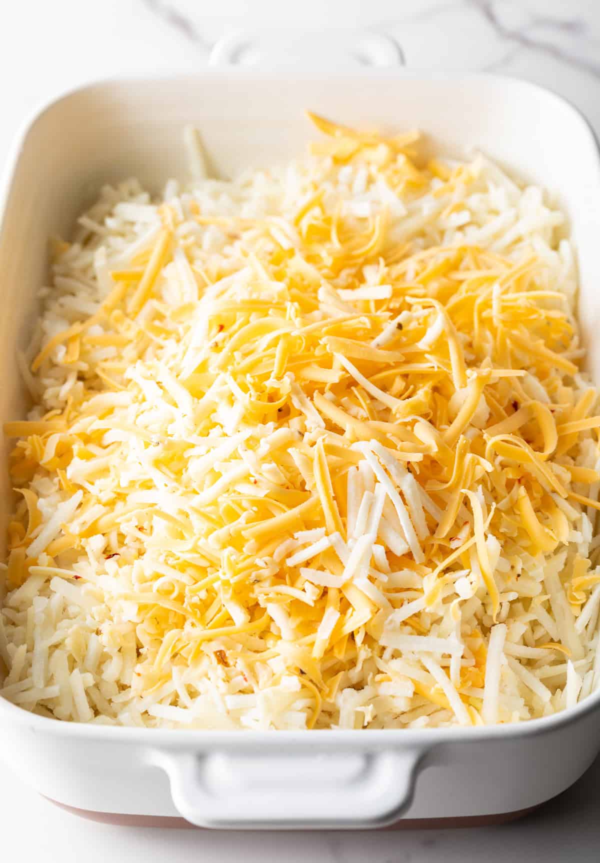 Shredded cheese is sitting on top frozen hash browns for a cheesy hashbrown casserole.