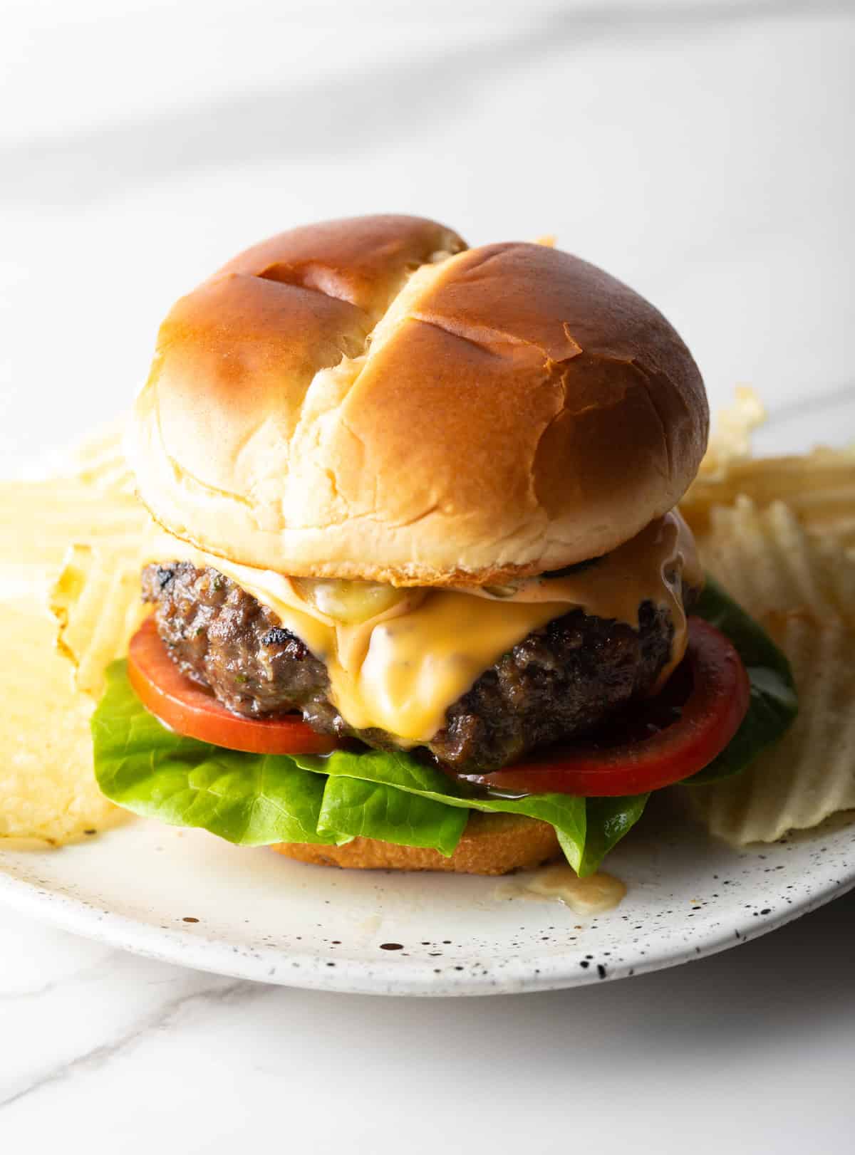 A juicy homemade crack burger topped with cheese, lettuce, and tomato, sitting on a white plate surrounded by chips.
