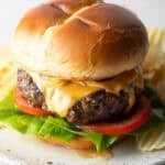 A juicy homemade crack burger topped with cheese, lettuce, and tomato, sitting on a white plate surrounded by chips.