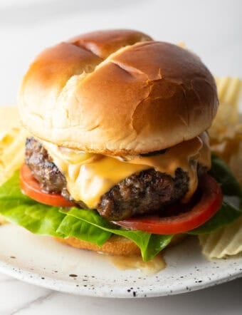 A juicy homemade crack burger topped with cheese, lettuce, and tomato, sitting on a white plate surrounded by chips.