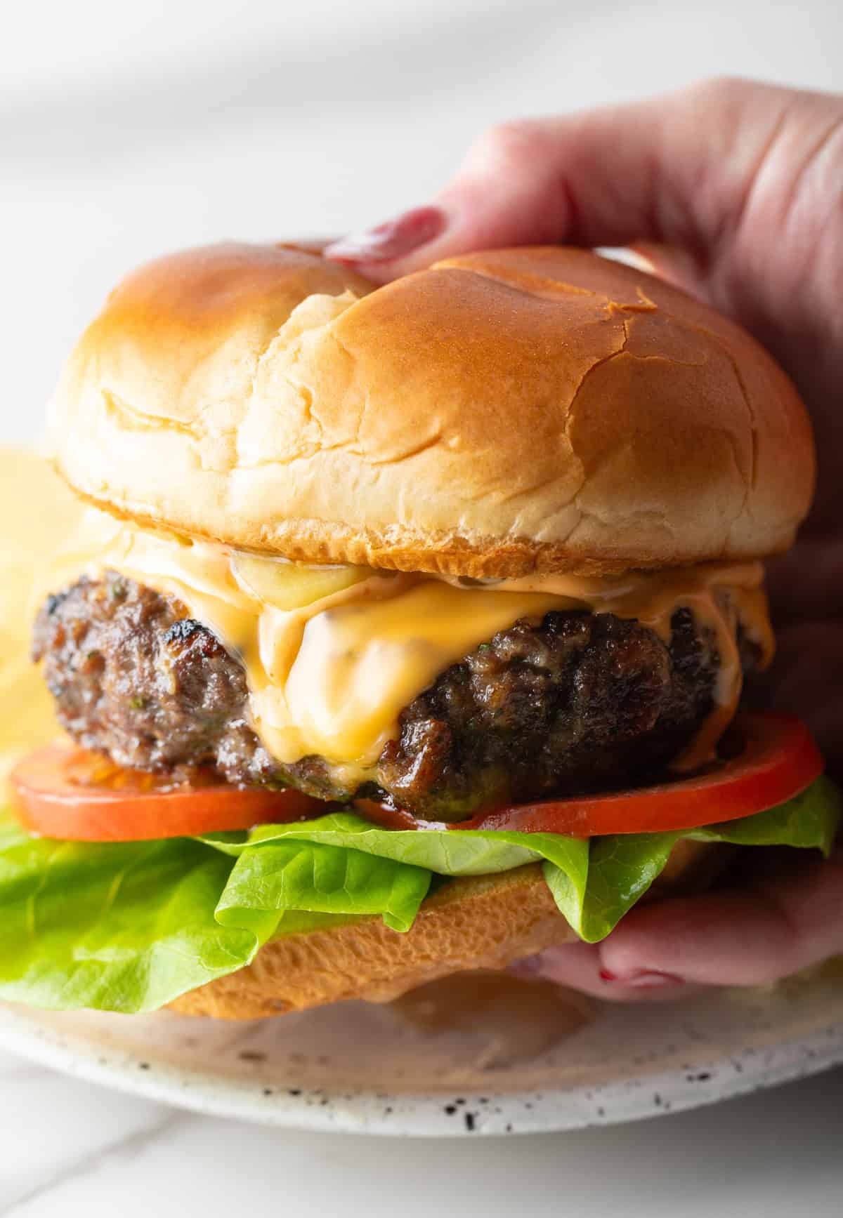 A hand is holding a cheese-topped crack burger over a white plate.
