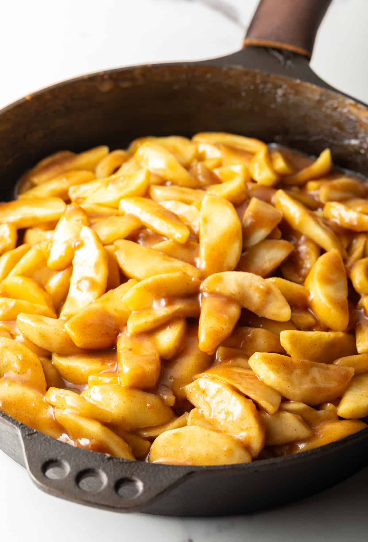 Sliced apples are being cooked in a black skillet with sugar and spices.