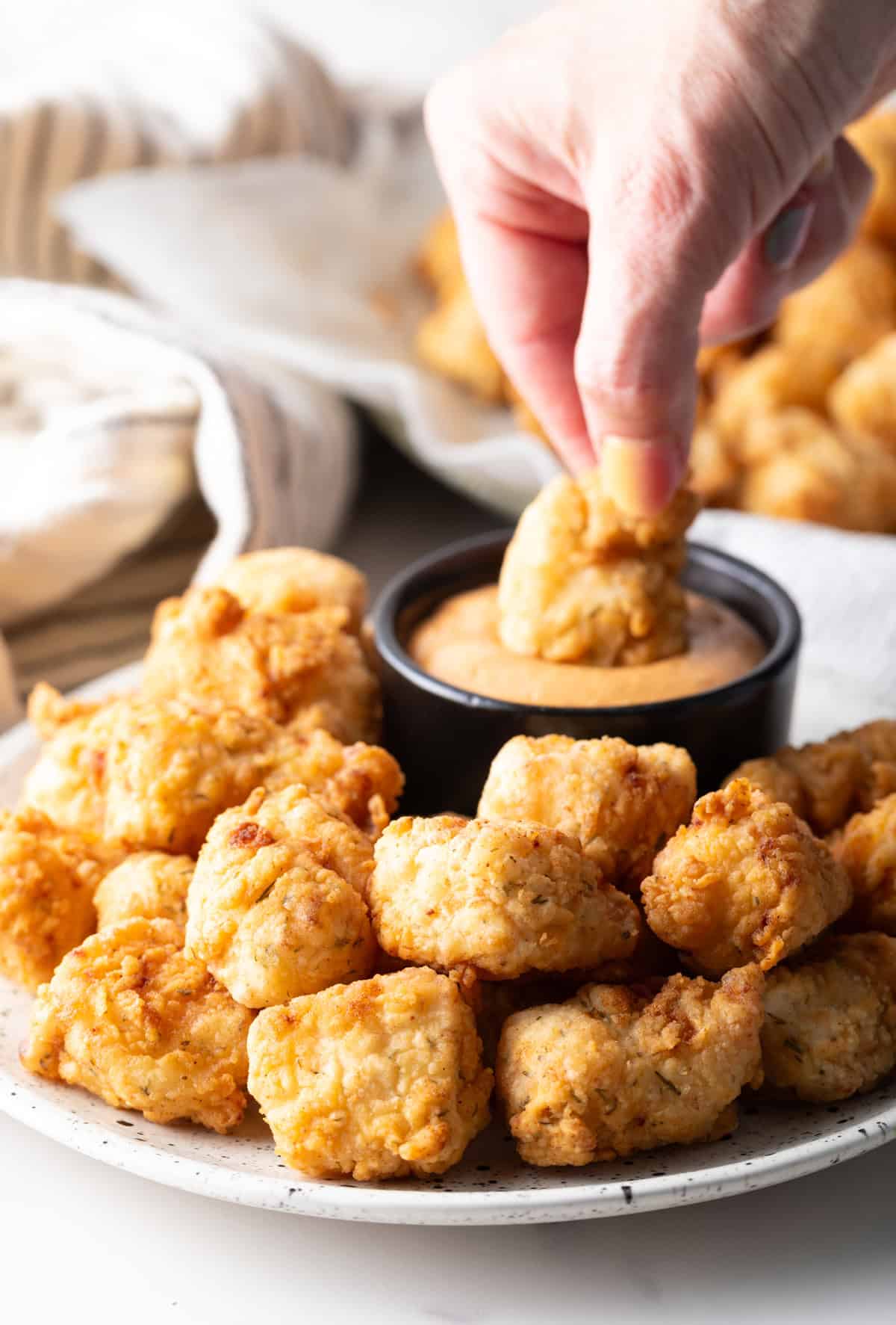 A hand is dunking a crispy homemade fish nugget into a ramekin of sauce.