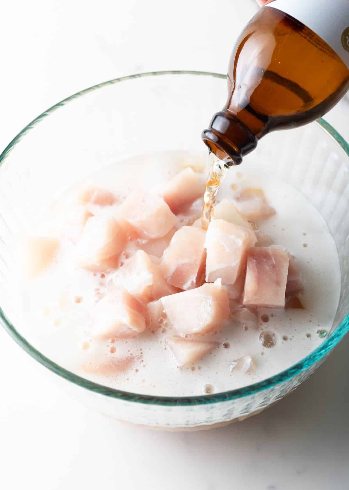 A bottle of beer is being poured into a glass bowl filled with raw chunks of white fish.