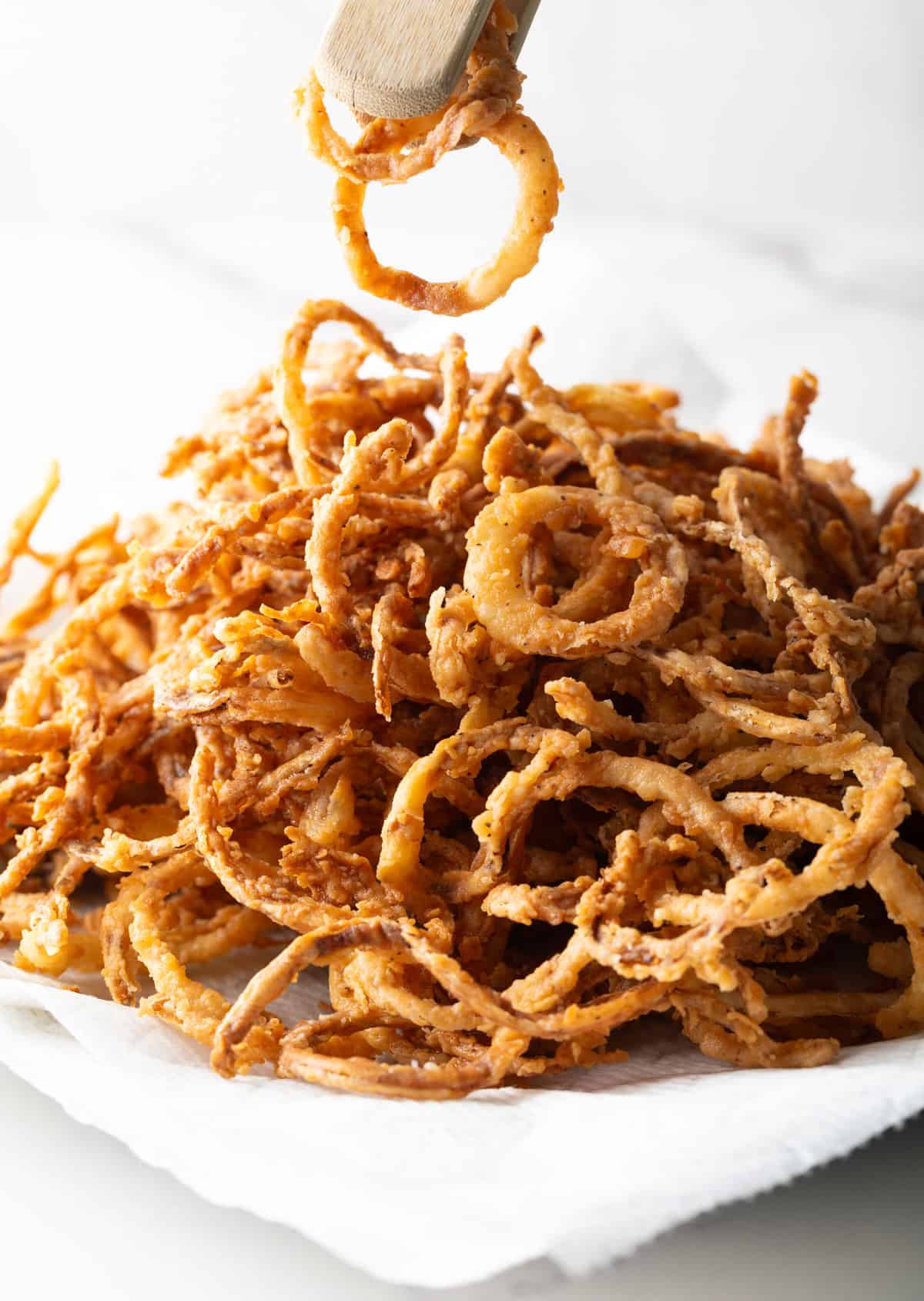 A pair of tongs is holding two French fried onions above a plate of the homemade French fried onions recipe.