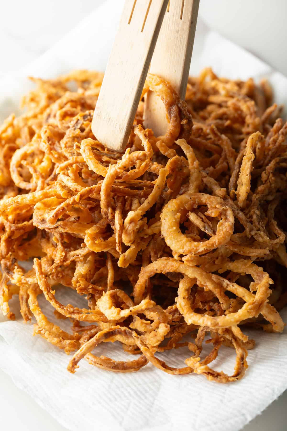 Homemade French fried onions are being added to a paper towel-lined plate with wooden tongs.