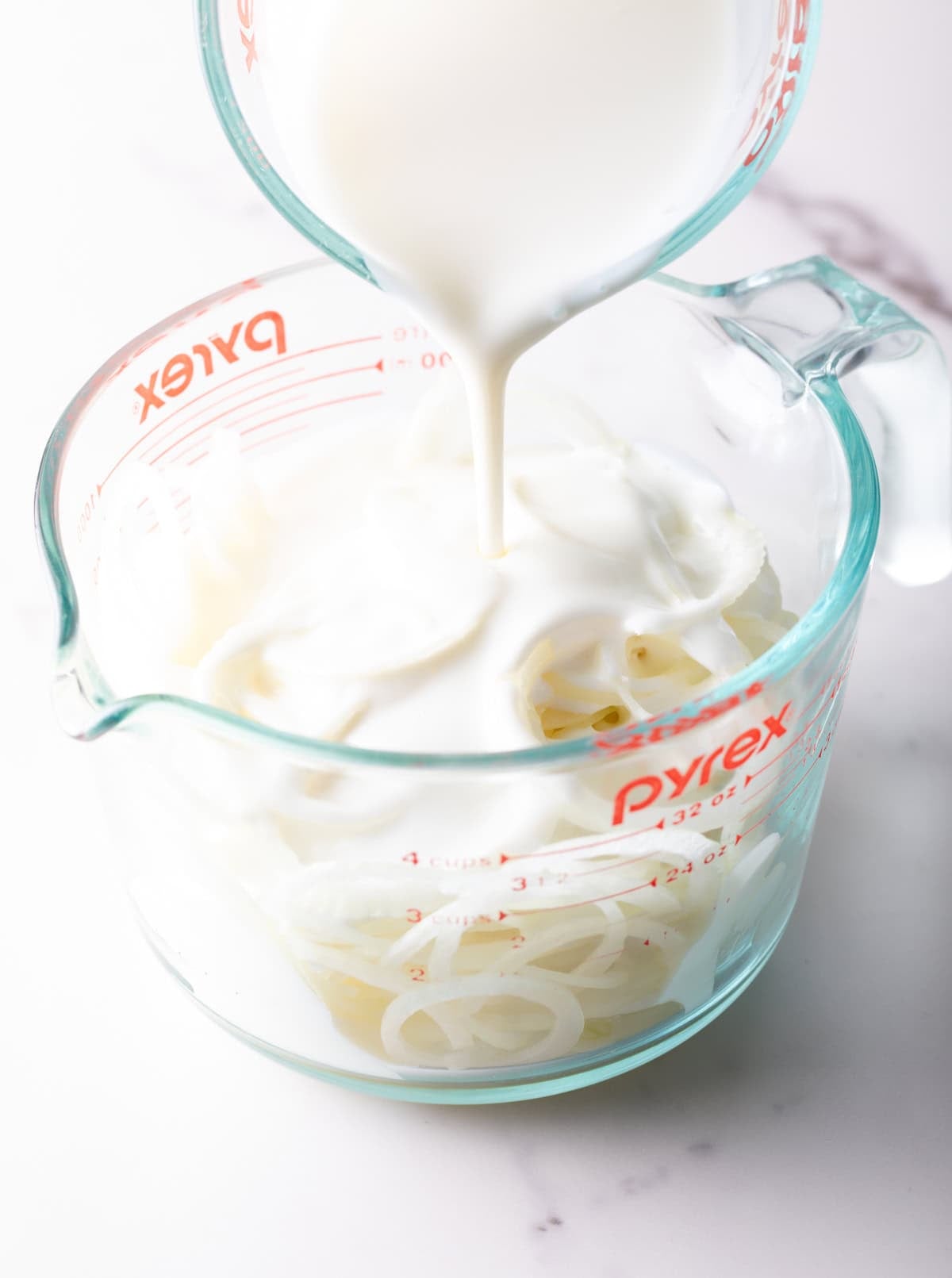 Buttermilk is being poured over onion slices that are sitting in a glass measuring pitcher.