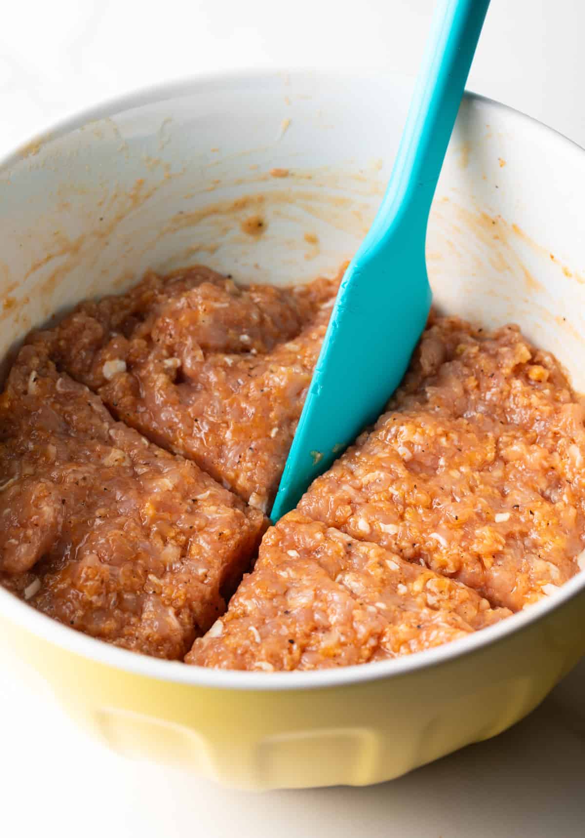 A ground chicken patties mixture in a white bowl, being divided into 4 portions with a blue spatula.