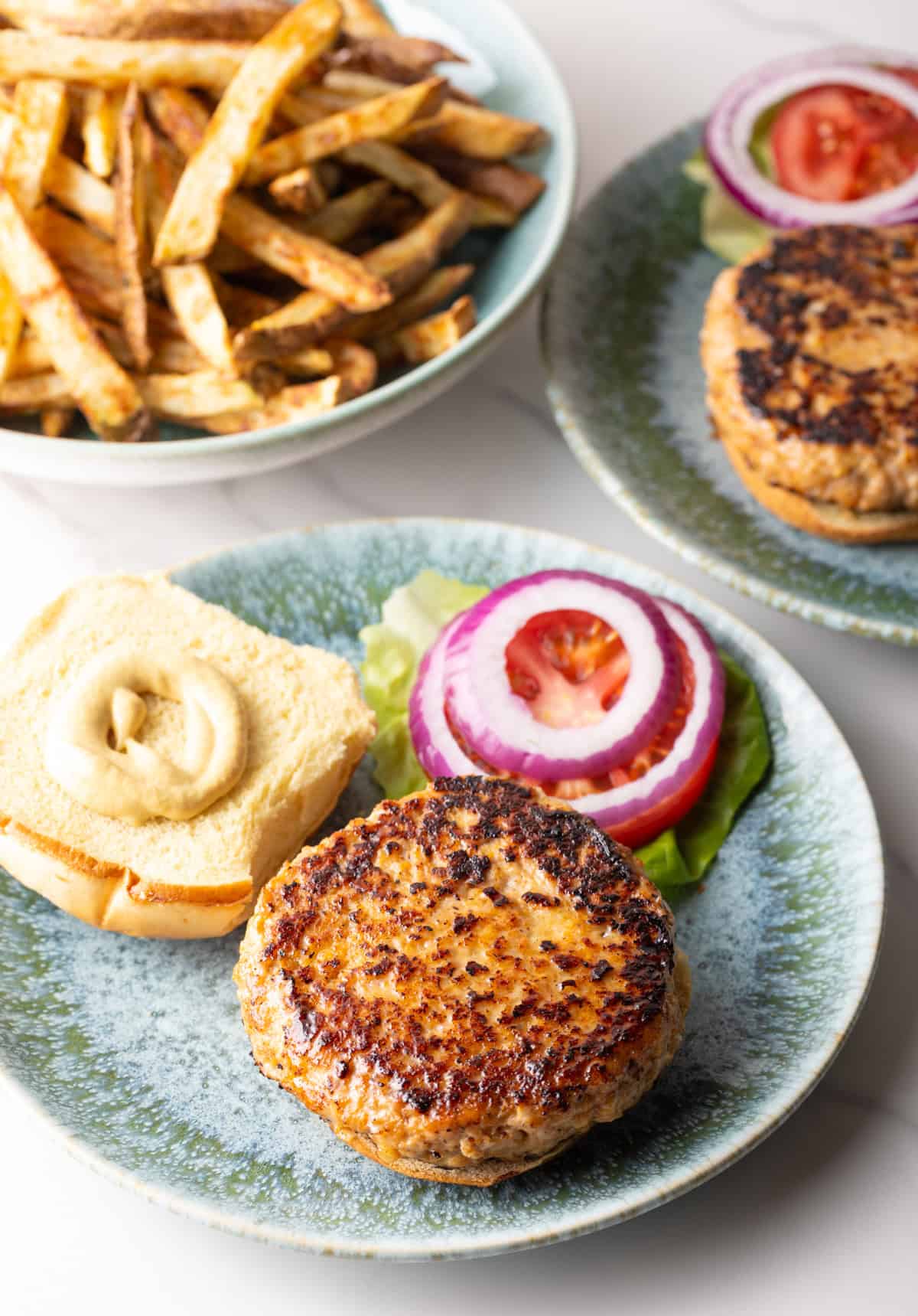 An open chicken patty sandwich with mayonnaise on half of the bun, sitting next to lettuce, tomatoes, and red onions.