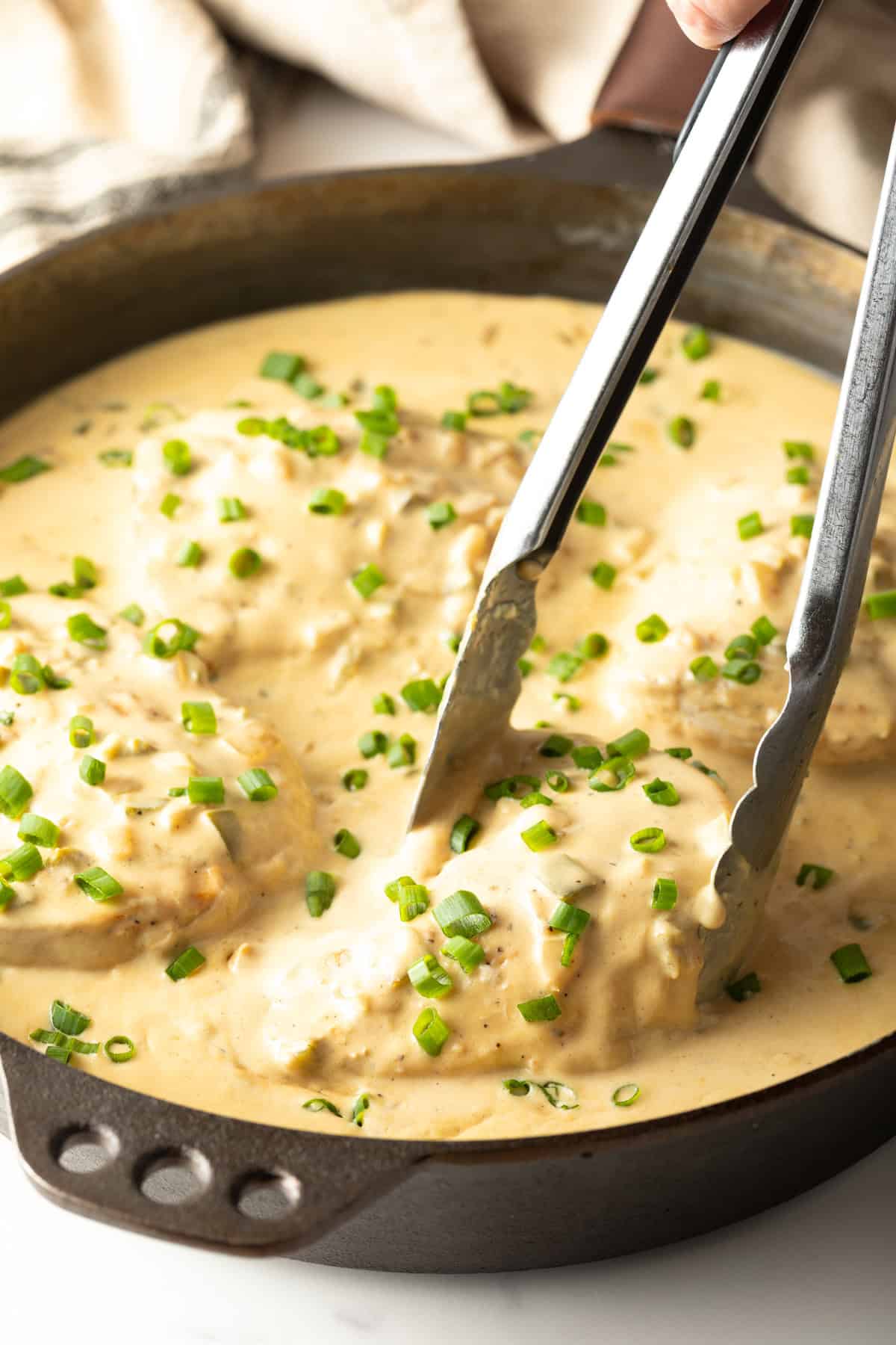 A pair of metal tongs is picking up a piece of jalapeno popper chicken from a black skillet.