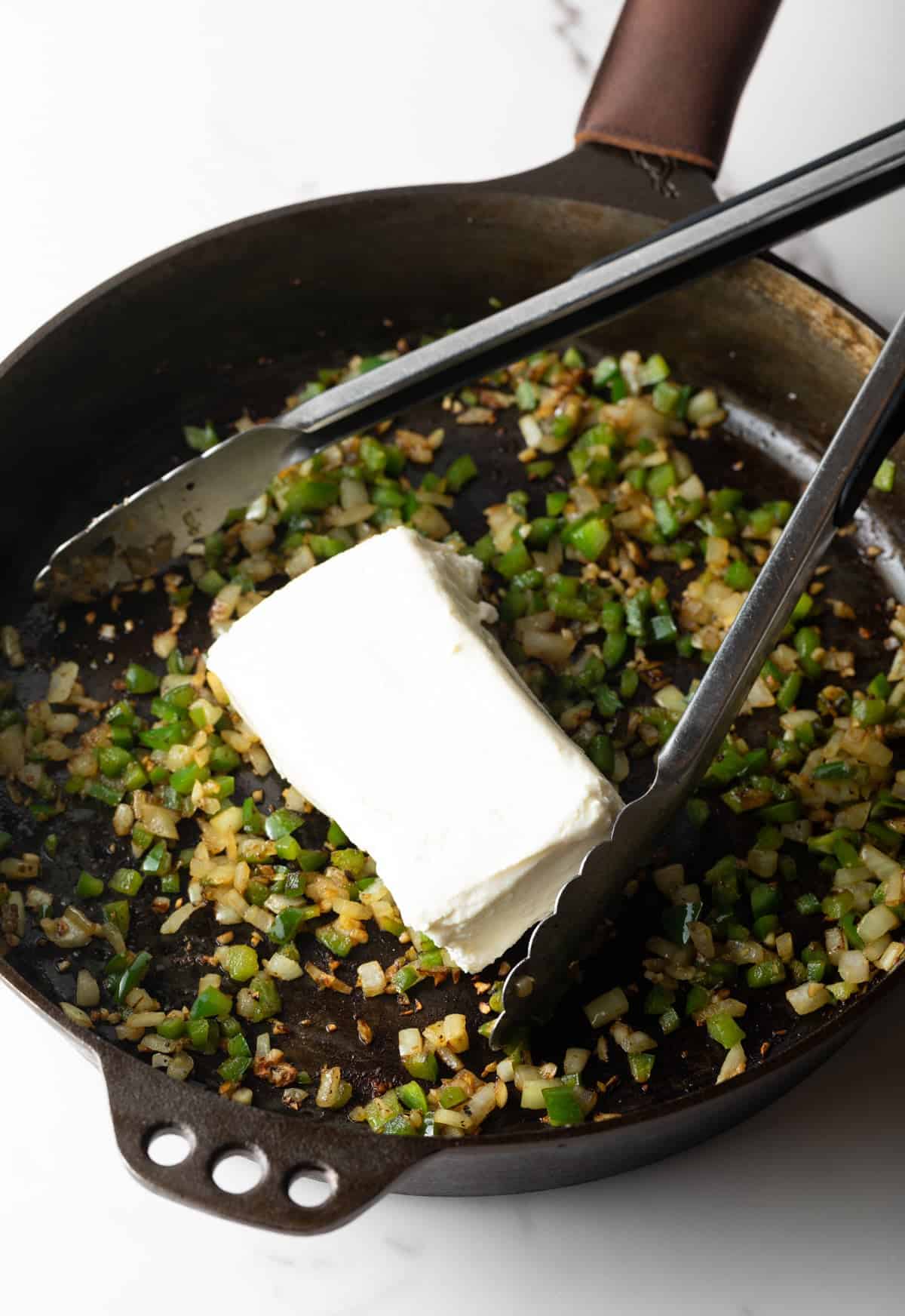 A block of cream cheese is being added to a sautéed veggie mixture in a black skillet.