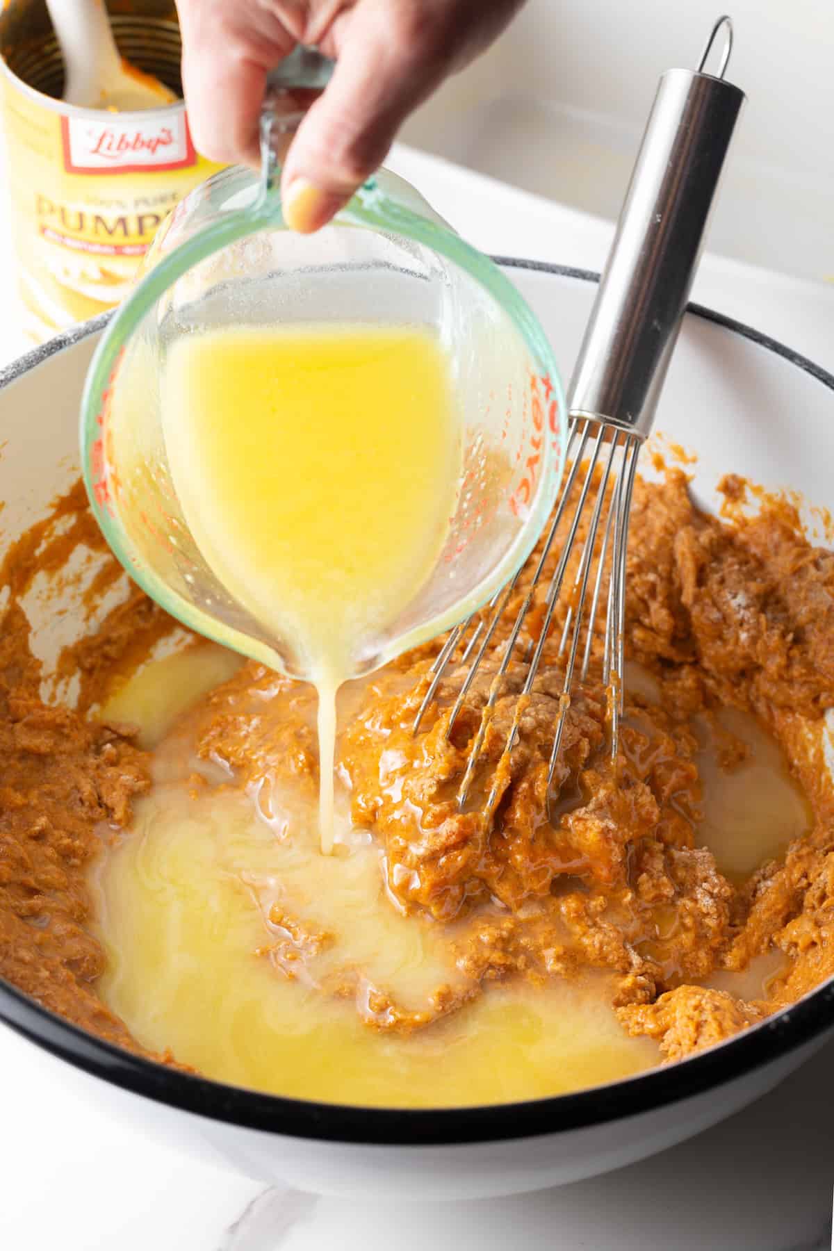 Butter is being poured over a pumpkin bread recipe mixture in a white bowl with a whisk.