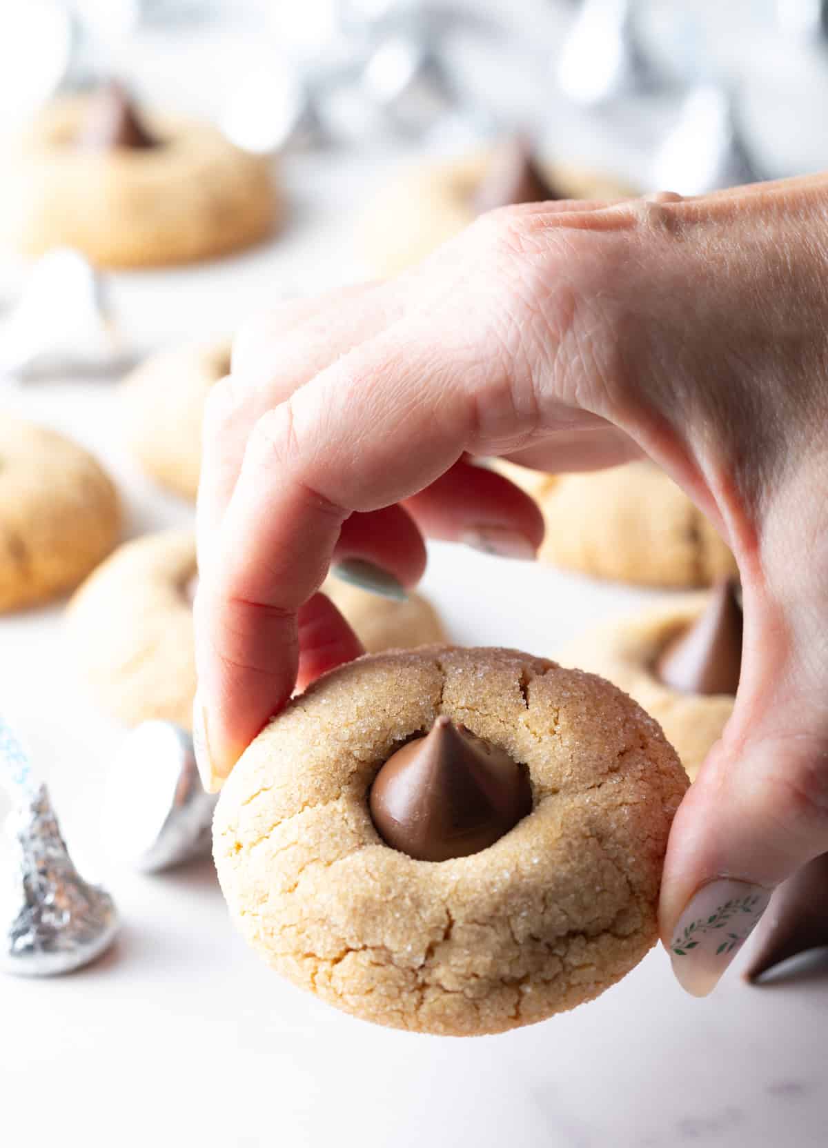 A hand holding a peanut butter kiss cookie to the camera above a tray of the cookies.