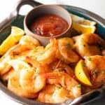 A bowl filled with a peel and eat shrimp recipe with cocktail sauce and lemon wedges on the side.