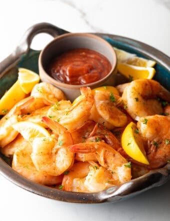 A bowl filled with a peel and eat shrimp recipe with cocktail sauce and lemon wedges on the side.