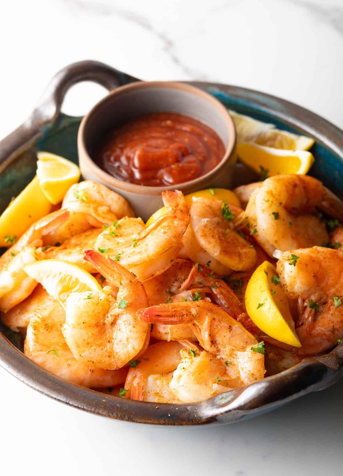 A bowl filled with a peel and eat shrimp recipe with cocktail sauce and lemon wedges on the side.