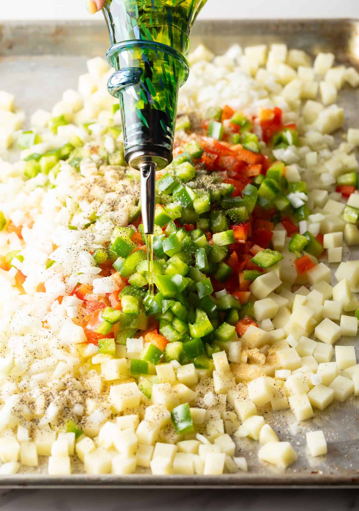Olive oil is being drizzled over diced potatoes, bell peppers, and onion on a baking sheet.