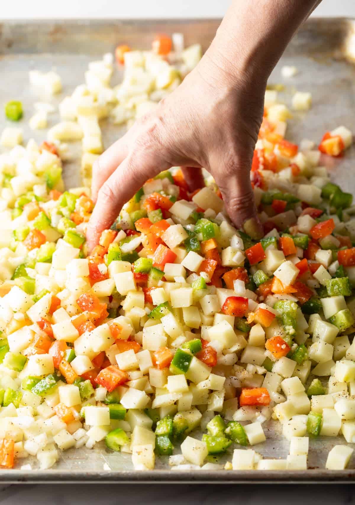 A hand is tossing the ingredients for a potatoes O'brien recipe with oil and seasonings on a baking sheet.