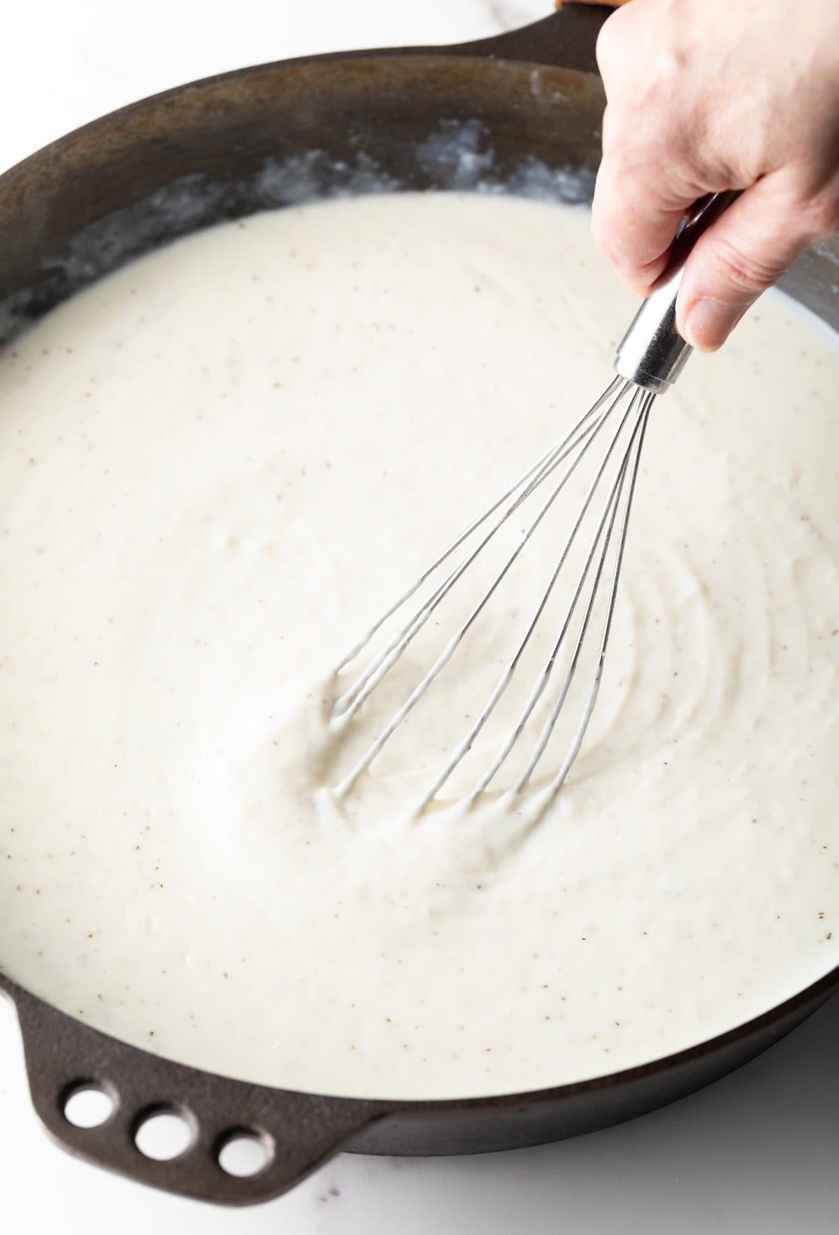 A roux for a white gravy recipe is being whisked in a black skillet.