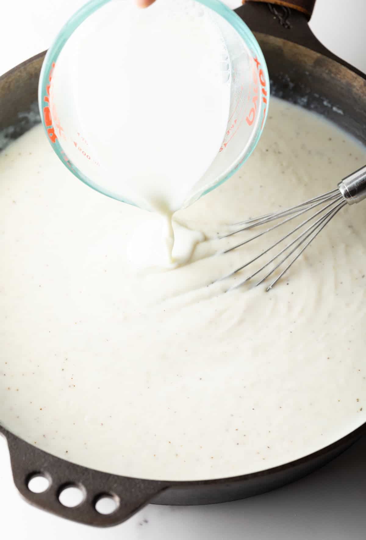 Milk is being poured into a black skillet for a white country gravy recipe.