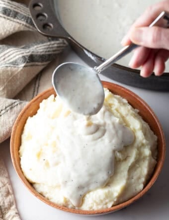 A white gravy recipe is being spooned over a bowl of mashed potatoes.