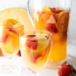 A pitcher and a wine glass filled with a white wine sangria recipe with fresh fruit floating in each.