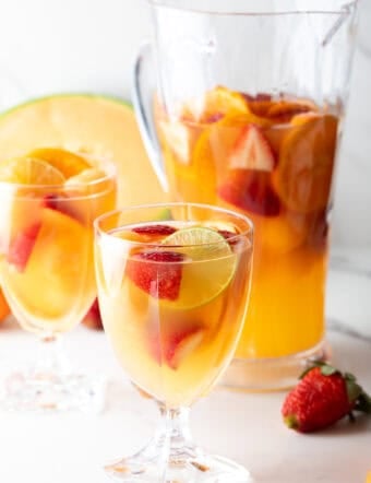 A pitcher and a wine glass filled with a white wine sangria recipe with fresh fruit floating in each.